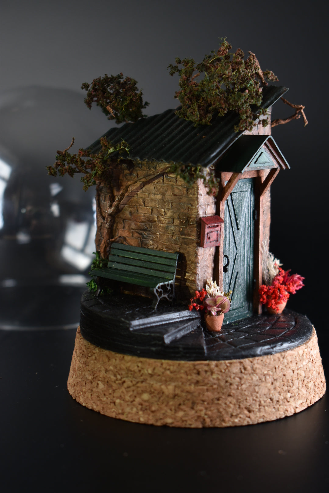 A miniature diorama in a glass dome, featuring the corner of an old brick house with a green door, awning, wrought-iron bench, and charming flower pots with floral arrangements. Totally handmade by HandiCraftArt