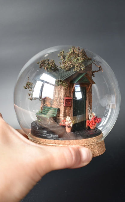 A miniature diorama in a glass dome, featuring the corner of an old brick house with a green door, awning, wrought-iron bench, and charming flower pots with floral arrangements. Totally handmade by HandiCraftArt