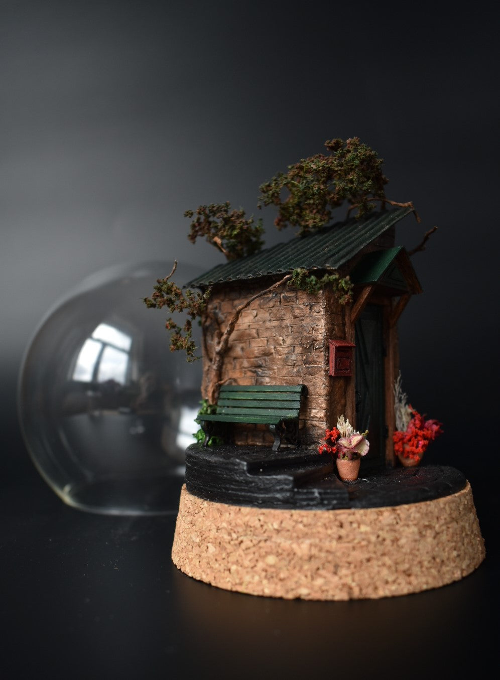 A miniature diorama in a glass dome, featuring the corner of an old brick house with a green door, awning, wrought-iron bench, and charming flower pots with floral arrangements. Totally handmade by HandiCraftArt