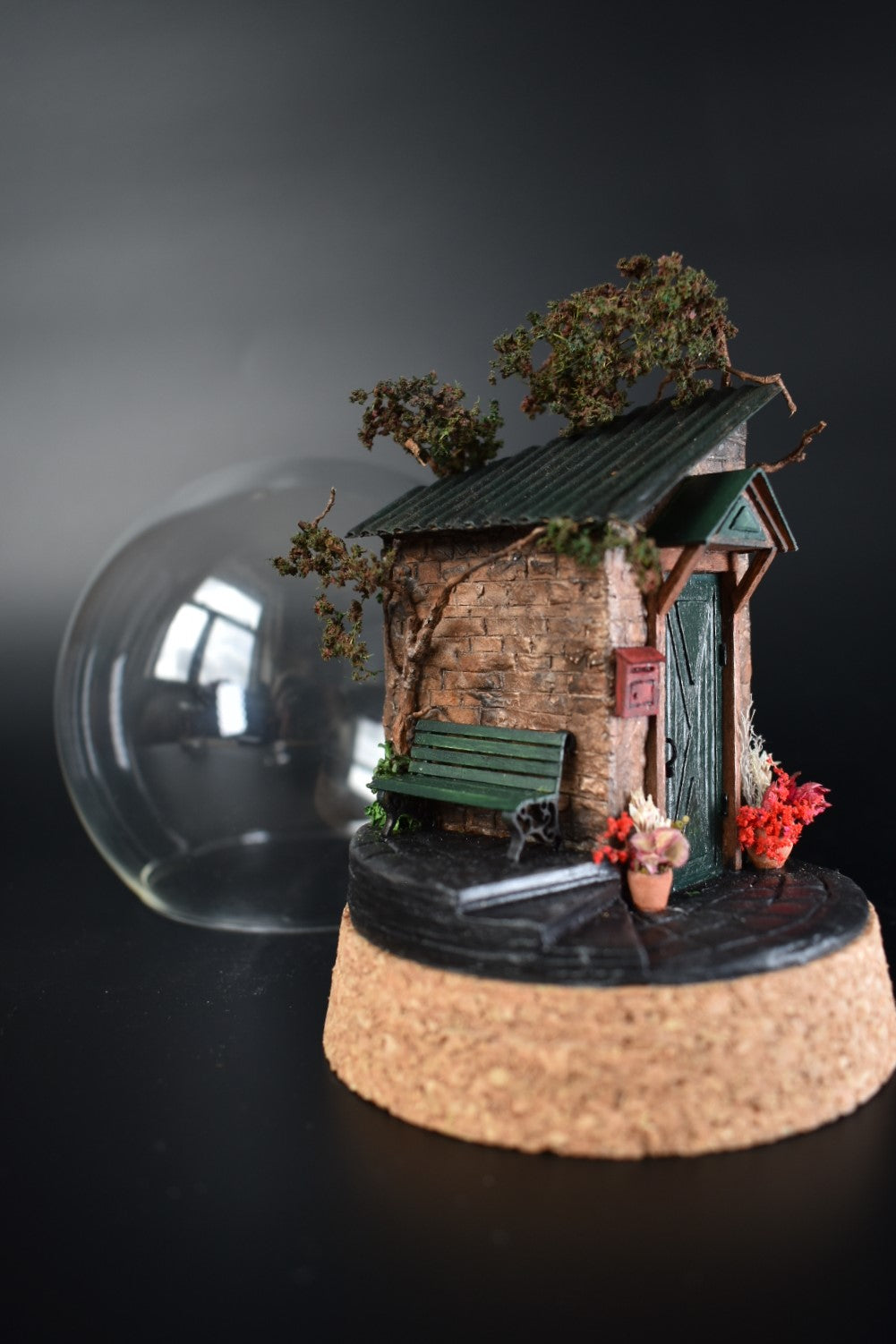 A miniature diorama in a glass dome, featuring the corner of an old brick house with a green door, awning, wrought-iron bench, and charming flower pots with floral arrangements. Totally handmade by HandiCraftArt