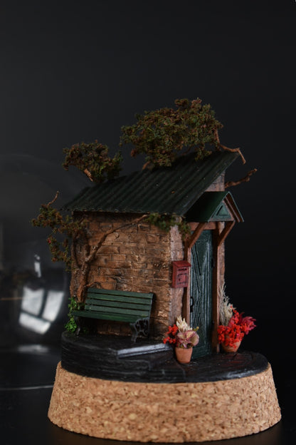 A miniature diorama in a glass dome, featuring the corner of an old brick house with a green door, awning, wrought-iron bench, and charming flower pots with floral arrangements. Totally handmade by HandiCraftArt