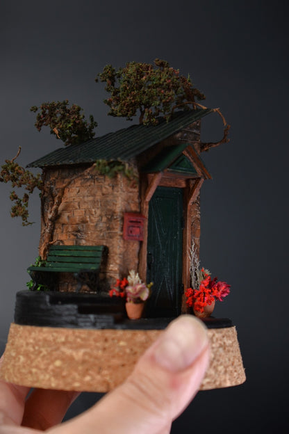 A miniature diorama in a glass dome, featuring the corner of an old brick house with a green door, awning, wrought-iron bench, and charming flower pots with floral arrangements.