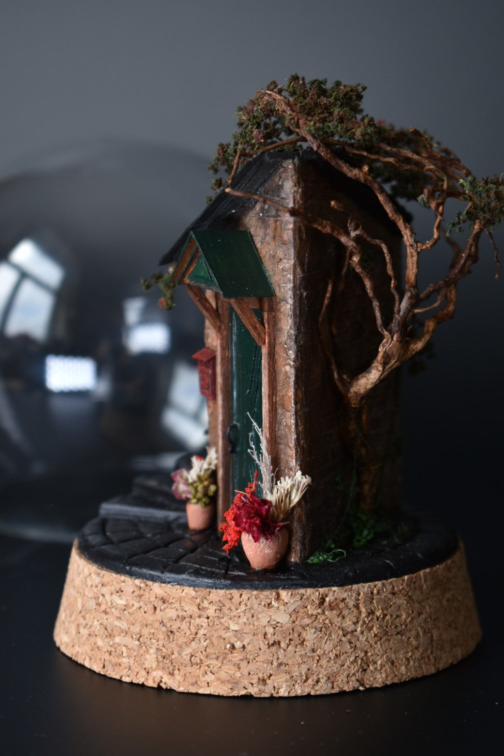 A miniature diorama in a glass dome, featuring the corner of an old brick house with a green door, awning, wrought-iron bench, and charming flower pots with floral arrangements. Totally handmade by HandiCraftArt