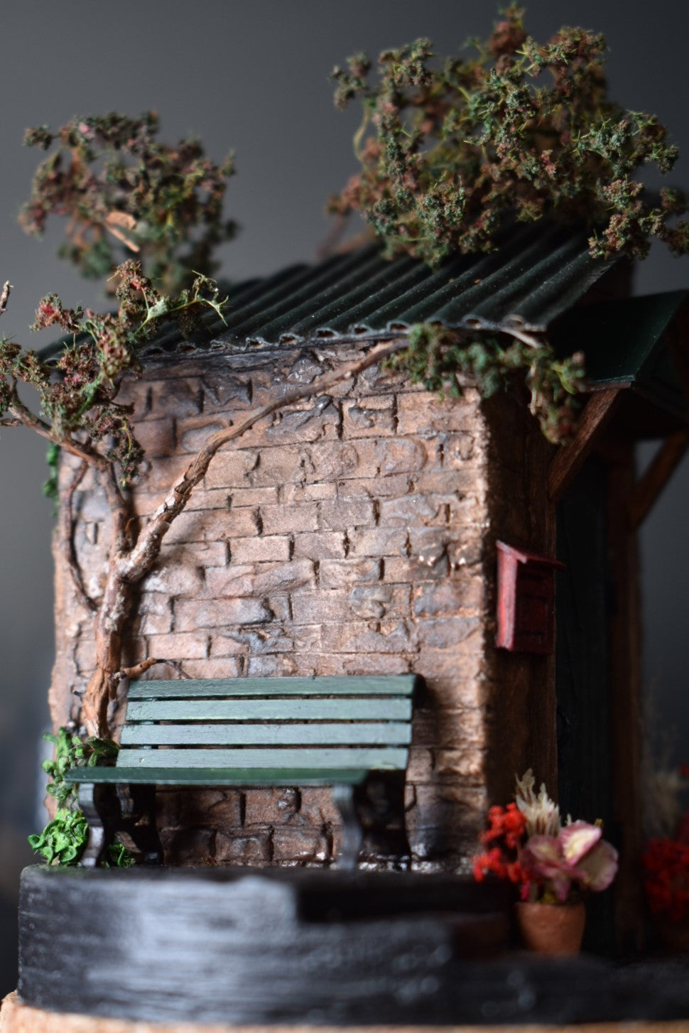 A miniature diorama in a glass dome, featuring the corner of an old brick house with a green door, awning, wrought-iron bench, and charming flower pots with floral arrangements. Totally handmade by HandiCraftArt