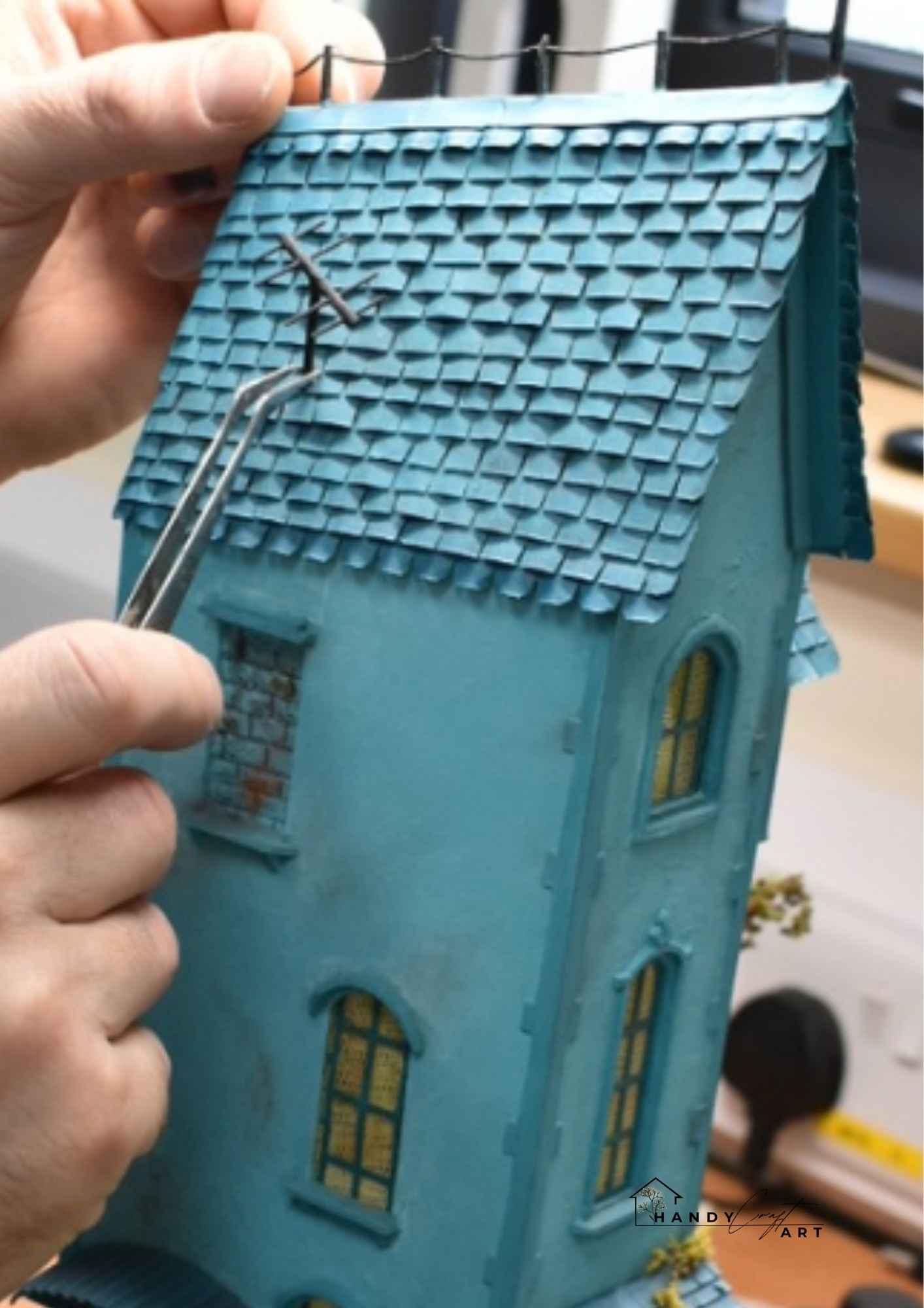 Handmade miniature diorama scene by HandiCraftArtIE. Teal model of a house with detailed roof shingles being adjusted by a hand.