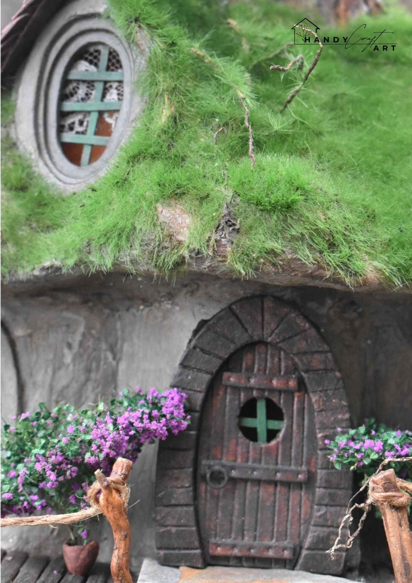 Handcrafted miniature fantasy diorama Gnome house with a wooden door and green roof, surrounded by purple flowers by HandicraftArtie. 