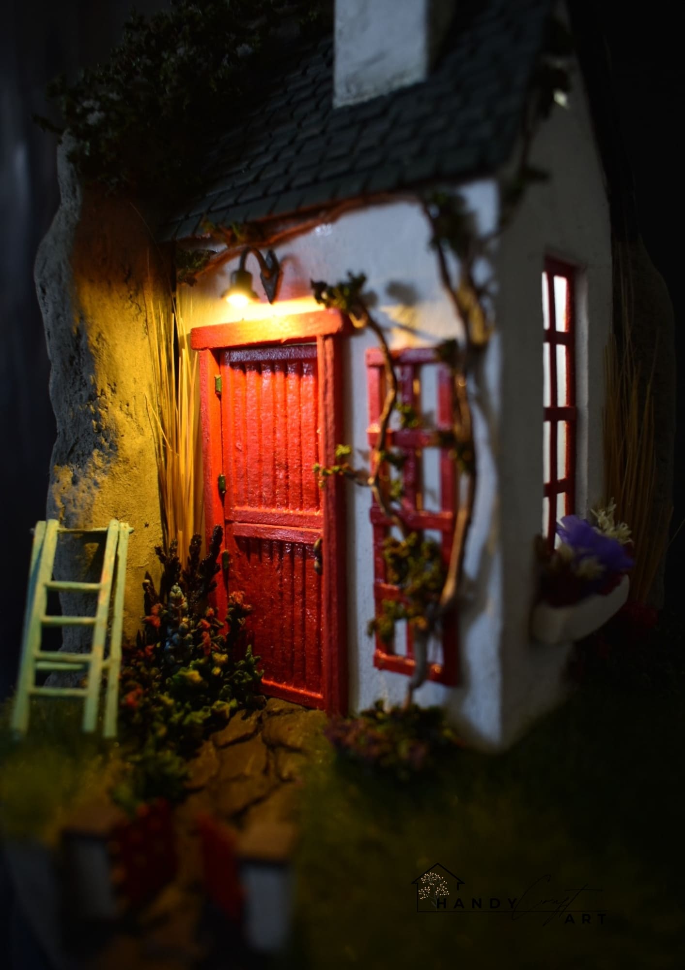 Small model house with a red door and white walls, illuminated from within and tiny ligtht.