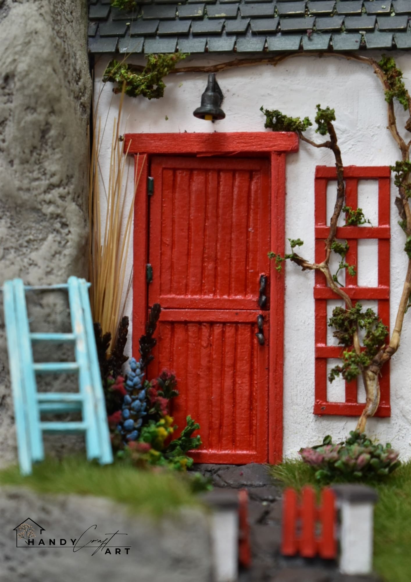 Miniature fairy house with a red door and white walls, featuring a small ladder and plants.