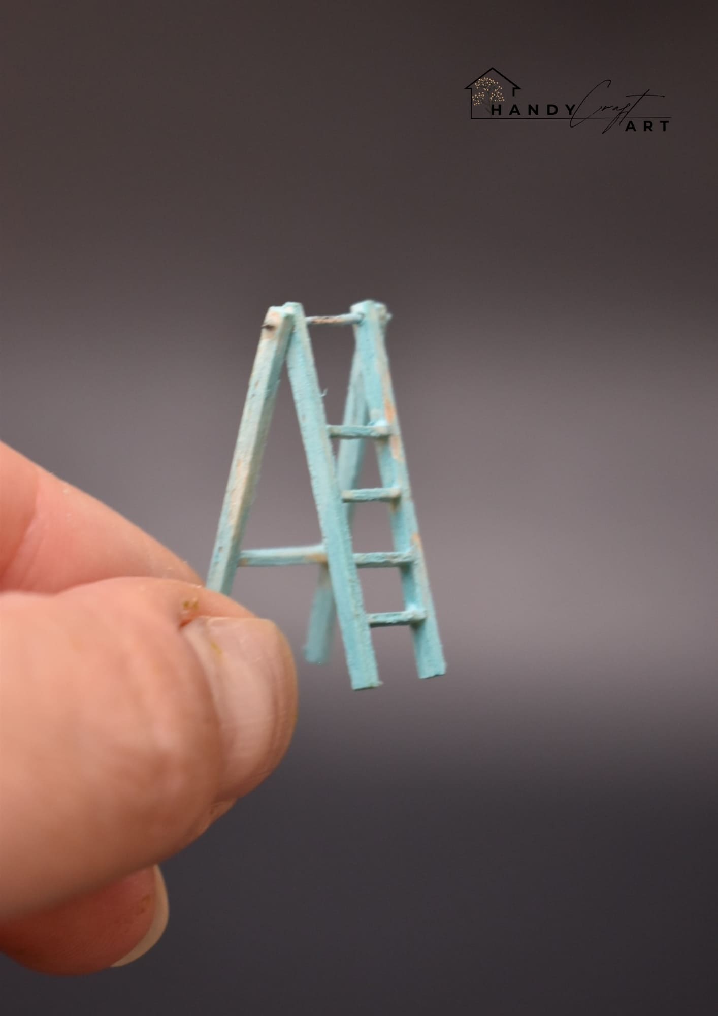 Handmade miniature blue ladder held by a hand against a gray background