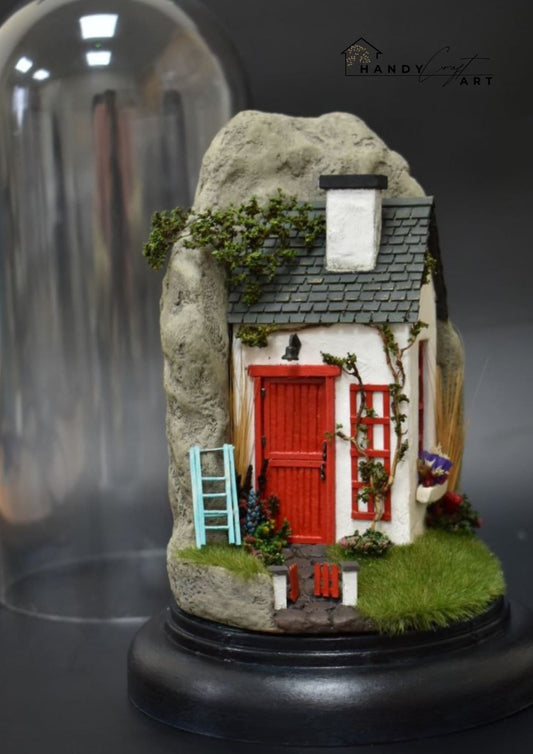 Miniature house with a red door on a rock, enclosed in a glass dome with HandyCraftArt branding.
