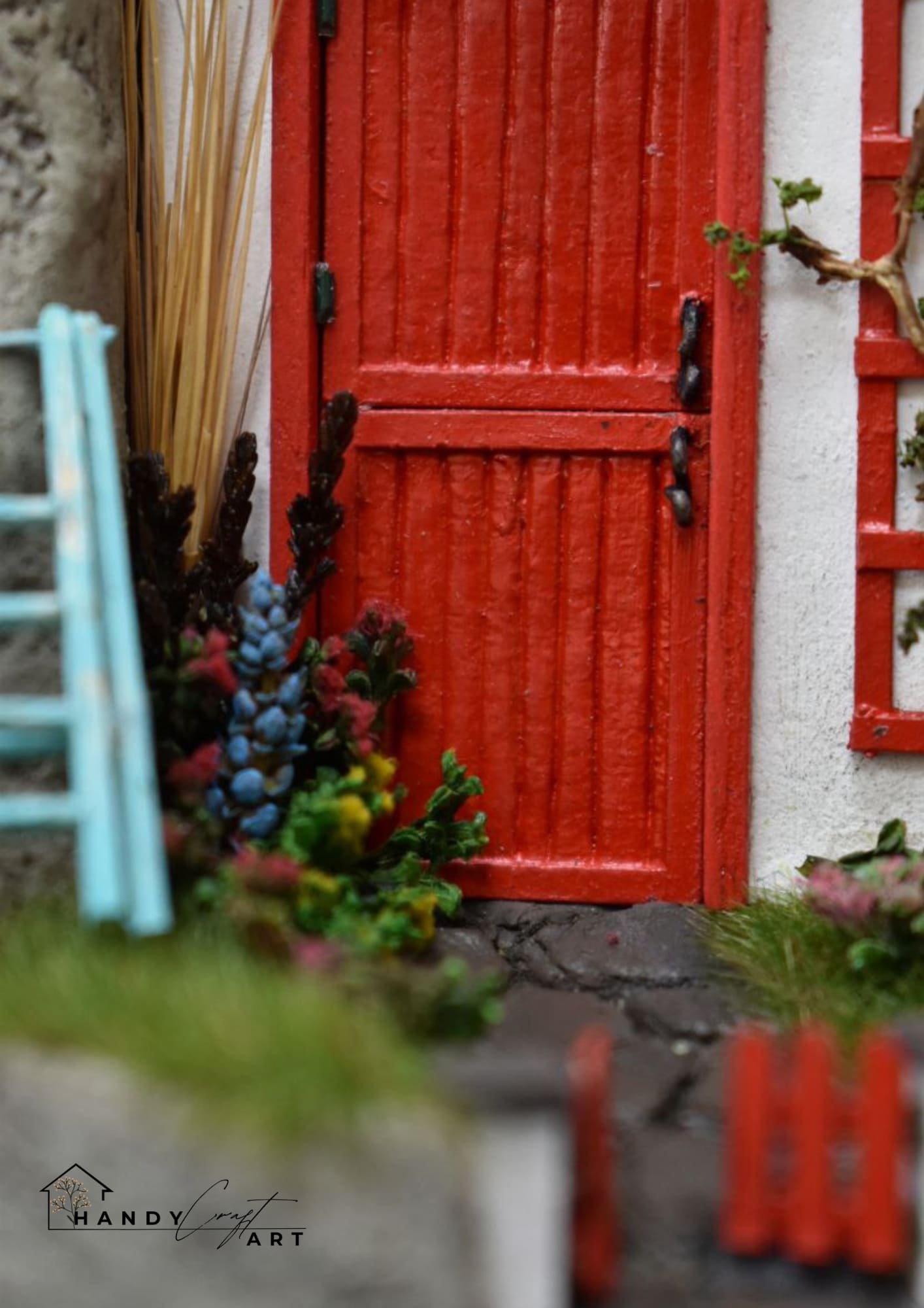 Miniature red door with plants and a ladder in front of a white wall. Handmade by HandiCraftArt